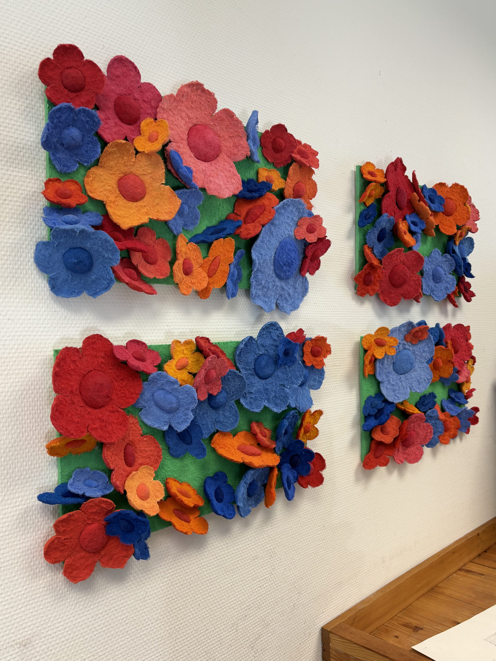 Vier Tafeln mit roten, blauen, rosa und orangefarbenen Blüten auf grünem Grund an einer weißen Wand.